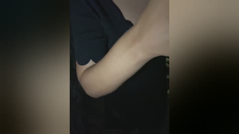 Media: Video of a person's arm and torso in a dark room, wearing a black shirt, with a blurred background.