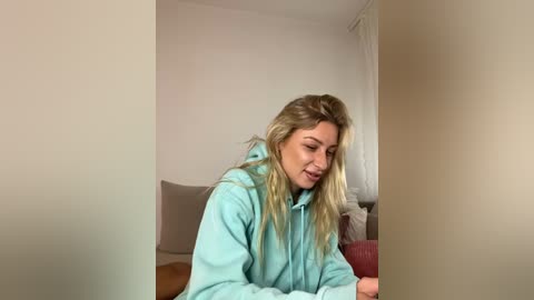 Media: A video of a young woman with long, blonde hair, wearing a light blue hoodie, sitting on a beige sofa in a minimalist, white-walled room.