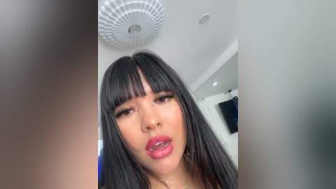 Media: Video of an Asian woman with long black hair and blunt bangs, wearing bright pink lipstick, looking seductively into the camera. Background includes a ceiling fan and part of a room with a TV.