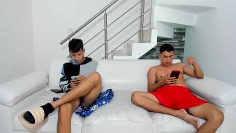 Media: Video of two shirtless young men, one in red shorts, the other in blue, sitting on a white leather couch, engrossed in their smartphones, in a modern, minimalist living room with white walls and a metal staircase.