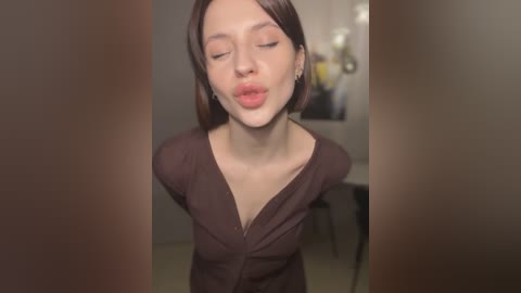 Media: Video of a young woman with light skin and straight, shoulder-length brown hair, wearing a dark brown V-neck top, puckering her lips in a playful manner. Background features a blurred, neutral-toned room with a framed artwork.