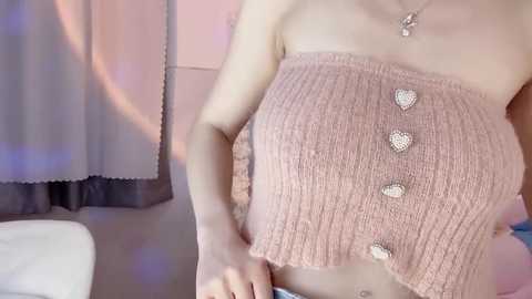 Video of a woman with fair skin wearing a pink, ribbed, strapless crop top adorned with heart-shaped buttons and a delicate necklace. Background features soft pastel curtains and a white couch.