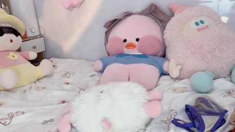 Video of a plush duck with pink feathers and a blue shirt, lying on a bed with white sheets patterned with small, colorful animals. Surrounding plush toys include a pink pig and a yellow duck.