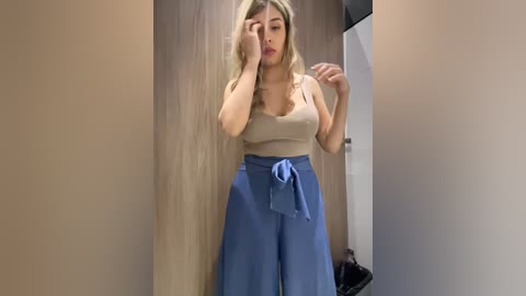 Video of a blonde woman in a beige tank top and high-waisted blue pants, standing in a wooden-walled room with a closed door behind her. She has her hands on her head and appears distressed.