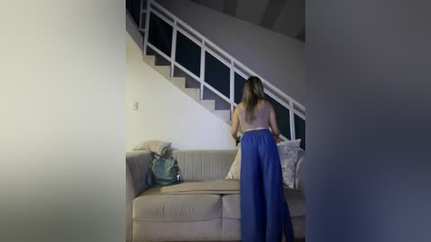 Video of a woman with long brown hair in a blue dress, walking away from a beige sofa with a green bag, in a modern, dimly lit room with a white staircase.