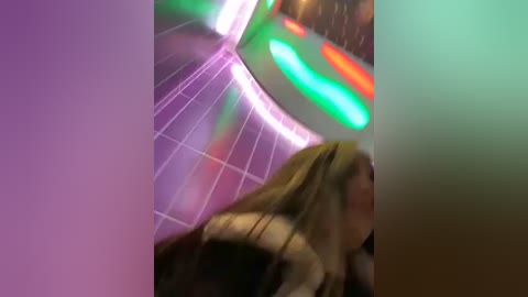 Media: A blurry video captures a woman with long, blonde hair, possibly Caucasian, wearing a dark jacket, walking in a neon-lit, retro-themed corridor with colorful, glowing signs.