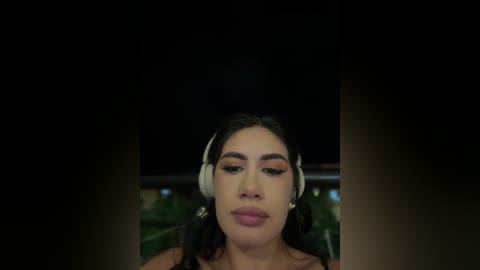 Media: Video of a Latina woman with dark hair, wearing headphones, looking serious. Background is dark, possibly night time with blurred greenery.