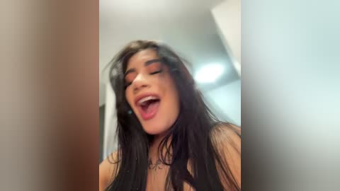 Media: A video of a young woman with long, dark hair, wearing a black top, captured in an indoor setting with blurred background. She has a joyful expression, mouth open, and eyes closed.