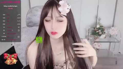 Media: Video of a pale-skinned woman with long dark hair, wearing a white flower headpiece, sitting in a gray chair, holding a black phone, in a minimalist room with a white wall and a glass table.