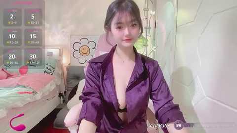 Media: A video of an Asian woman with fair skin, dark hair, and a purple satin robe, sitting on a bed with a pink and white bedspread, in a dimly lit, modern room with a floral wall art.