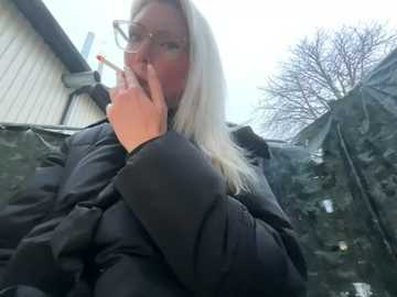 Media: Video of a blonde woman with glasses, wearing a black puffy coat, smoking a cigarette in a dark, tarp-covered outdoor setting.