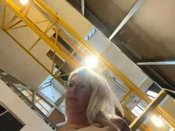 Media: Video of a blonde woman in glasses, wearing a beige top, standing in a modern, industrial space with yellow beams and exposed metal.