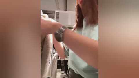 Media: A blurry video shows a woman with light skin and brown hair, wearing a light blue shirt, ironing a beige shirt. The background includes a white ironing board and a white microwave.