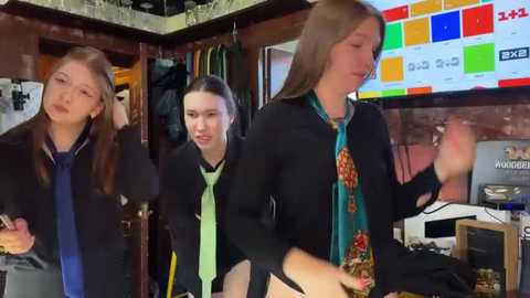 Media: Video of four young women in a cozy, cluttered room with wooden walls, one wearing a black cardigan and green tie, another a black jacket, holding a phone.