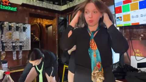 Media: Video of a woman with long brown hair, wearing a black blazer and colorful scarf, adjusting her hair in a pub with wooden decor, bar signs, and a TV screen.