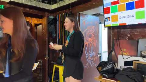 Media: Video of a young woman with long brown hair, wearing a black shirt and shorts, playing a video game in a cozy, dimly lit room with colorful graffiti on the wall and a flat-screen TV displaying game-related content.