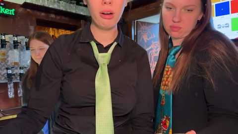 Media: Video of two women in a pub, one in a black shirt and green tie, the other in a colorful jacket, standing by a bar with a green sign, shelves, and a glass shelf.