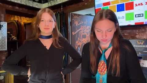 Media: Video of two young women in a bar, one wearing a black shirt and choker, the other in a black top and colorful tie. Background includes a colorful sports poster and graffiti on a wall.