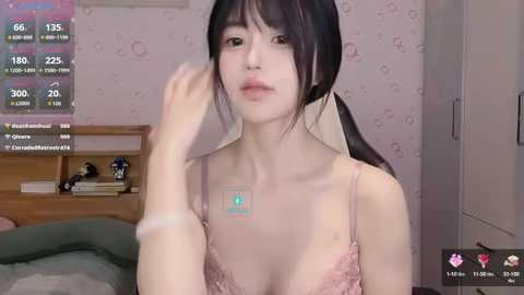 Media: A young Asian woman with pale skin and long black hair, wearing a pink lace bra, sits in a bedroom with pink wallpaper. Her face is neutral, and a digital health app is displayed on her chest.