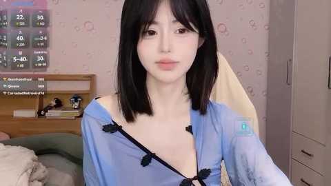 Media: Video of a young East Asian woman with fair skin, straight black hair, and a slender build, wearing a blue robe with a black bow. Background shows a cluttered bedroom with a bed, wall clock, and a TV.