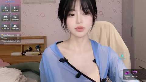 Media: Video of a young Asian woman with pale skin, black hair, and a petite frame, wearing a light blue, see-through robe, sitting in a dimly lit room with a TV displaying weather information.