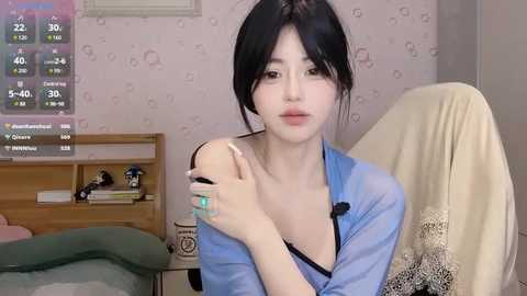 Media: Video of an Asian woman with fair skin, black hair, wearing a blue shirt, sitting in a beige chair, holding a drink, in a pastel-patterned room.