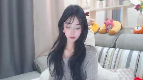 Media: A video of a young woman with long black hair, wearing a grey sweater, sitting on a light grey couch in a modern living room with striped pillows, plush toys, and potted plants.