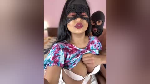 Media: Video of a woman in a black mask with pink lipstick, wearing a tie-dye shirt, exposing her large breasts. A man in a black mask holds her from behind. Background is a pink wall and bed.