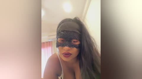 Media: Video of a woman with long black hair, wearing a black lace mask, and a gray tank top, standing indoors with blurred background.