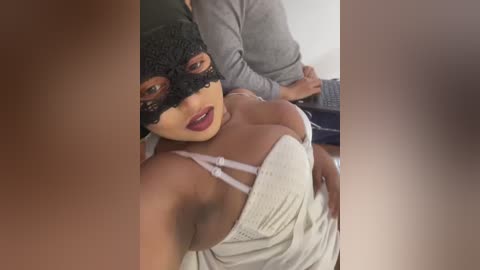 Media: Video of a dark-skinned woman with a black lace mask, red lipstick, and a white spaghetti strap top, partially exposing her breasts. A man's hand is visible on her shoulder, in a dimly lit room.