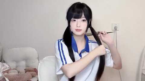 Media: Video of an East Asian woman with long black hair, wearing a white polo shirt with blue stripes, fixing her hair in a bedroom with a white tufted headboard and beige walls.