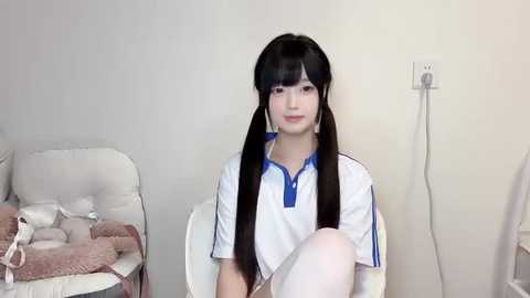 Media: A video of a young woman with long black pigtails, wearing a white sports jersey with blue trim, seated on a white chair in a minimalist room with a beige wall and a teddy bear.