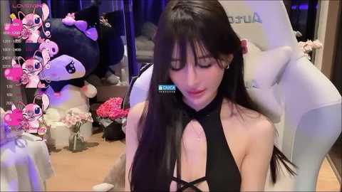 Media: A screenshot from a live stream shows a fair-skinned woman with long, straight black hair, wearing a black halterneck top, sitting on a white gaming chair. The background features plush toys and pink flowers.