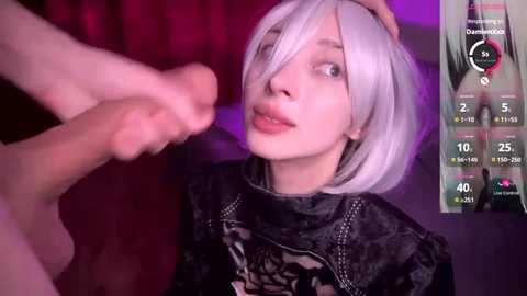 Media: A video of a light-skinned woman with platinum blonde hair in a bob cut, wearing a black lace top, playfully holding a man's foot. Background features a red curtain and a virtual reality interface.