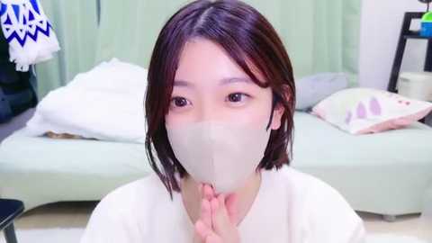 Media: Video of a young Asian woman with shoulder-length brown hair, wearing a white mask and a white top, kneeling in a pastel-colored room with a bed, pillows, and a wall-mounted shelf.