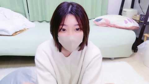 Media: Video of an Asian woman with short dark hair, wearing a white mask and long-sleeved shirt, kneeling on a white rug in a minimalist, pastel-colored bedroom with a bed and green curtains.