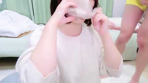 Media: Video of a fair-skinned woman with short dark hair, wearing a white sweater, playfully biting her own arm, in a light-colored room with a bed, green curtains, and yellow shorts in the background.