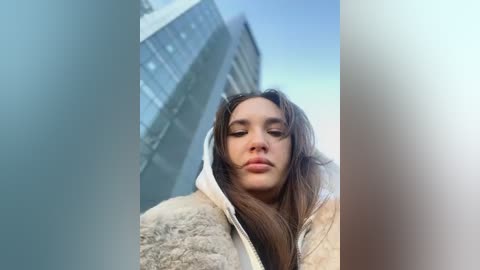 Media: Video of a young woman with long brown hair, wearing a beige fur coat, standing in front of a modern building with large windows, under a clear blue sky.