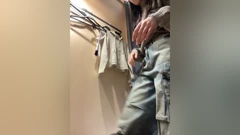 Media: Video of a person in a cramped, beige-walled closet with hanging clothes. They wear a grey sweater and blue jeans, reaching for a hanger, with a blurred background.