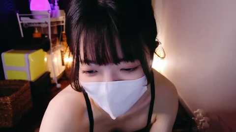 Media: Video of a young Asian woman with black hair and bangs, wearing a white face mask and black top, kneeling in a dimly lit room with a yellow box and a lamp.