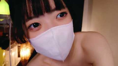 Media: A close-up video of an Asian woman with straight black hair and blue eyes, wearing a white surgical mask, indoors with a warm, dimly lit background.