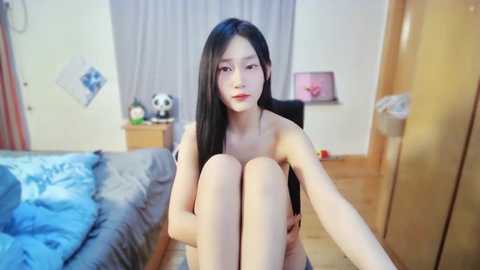 Media: Video of an East Asian woman with long black hair, sitting nude on a bed with a blue comforter, in a modest bedroom with wooden furniture and a panda figurine.