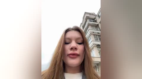 Media: Video of a young woman with long, straight, light brown hair, fair skin, and a neutral expression, standing in front of an urban background with a multi-story building in the background.