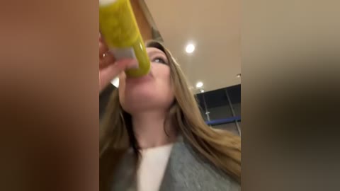 Media: A blurry video of a young woman with long, light brown hair, wearing a grey jacket, drinking a yellow beverage from a tall glass bottle, in a modern, brightly-lit room with a wooden floor and glass doors in the background.