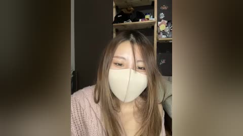 Media: Video of an East Asian woman with long brown hair, wearing a beige mask and a pink knit sweater, sitting in a dimly lit room with a wooden shelf behind her.