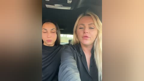 Media: Video of a blonde woman with a serene expression and a dark-haired man in a car, both wearing black clothing. The background shows the car interior.