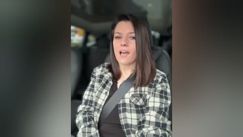 A video of a young woman with shoulder-length brown hair, wearing a black shirt and a plaid flannel, seated in a car with a seatbelt on, looking forward, mouth slightly open, blurred background.