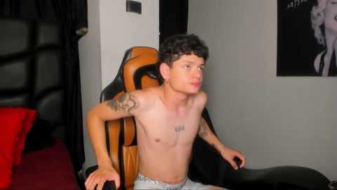 Media: Video of a shirtless young man with curly black hair, tattoos, and \"HYPE\" on his chest, wearing grey pajama pants, sitting in a gaming chair, in a dimly lit room with a red carpet.