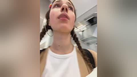 Media: Video of a young woman with brown braids, wearing a white shirt and beige cardigan, standing in an airport with blurred background and overhead lighting.