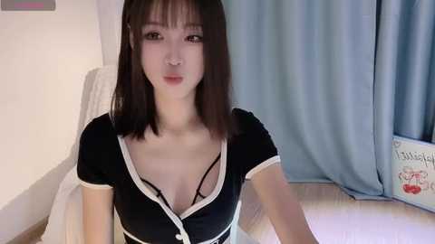 Media: Video of a young Asian woman with straight black hair, wearing a black maid costume with a plunging neckline, sitting on a bed in a room with light blue curtains.